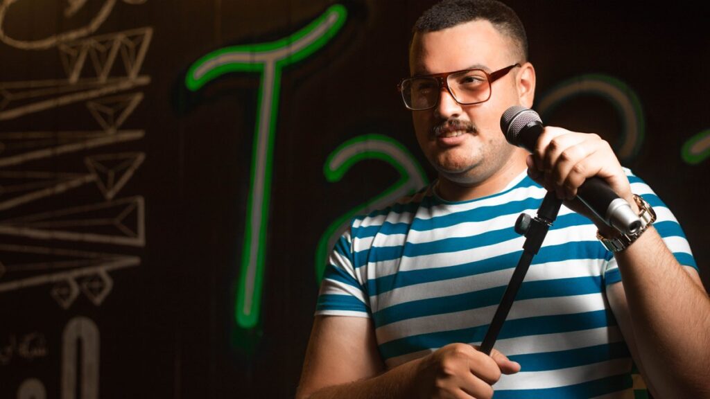 Man holding a microphone doing stand-up comedy in casual striped shirt on stage.